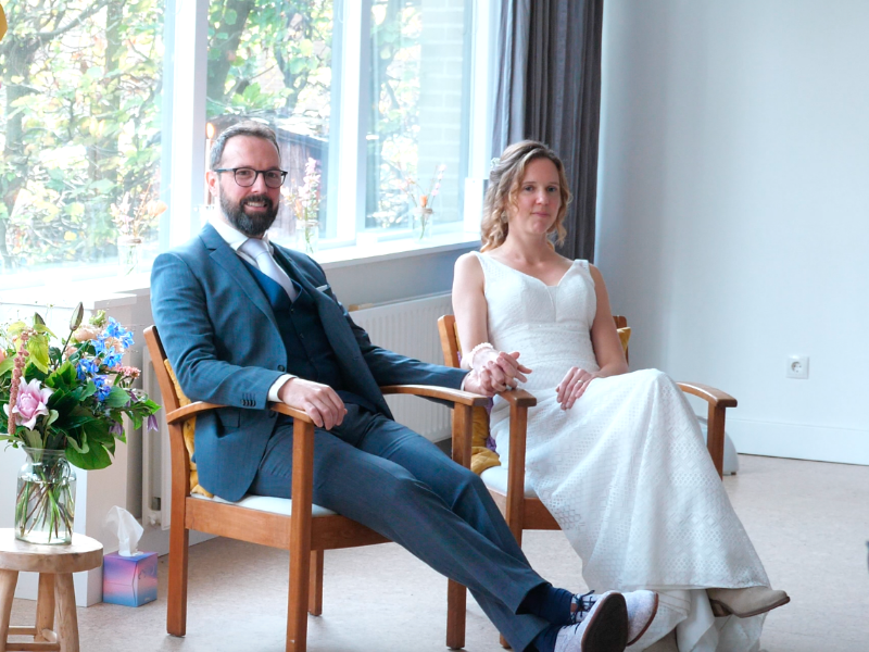 Bruidspaar Bianca en Paul zitten ieder in een stoel. Paul draagt een lichtblauw pak en Bianca een lange witte jurk. Zij houden elkaars hand vast en glimlachen.