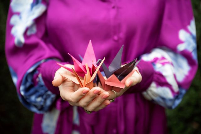 Tolk van Hartzaken Linda Blij houdt haar handen naar voren. In haar handen liggen origami kraanvogels in verschillende kleuren die zij zelf heeft gevouwen.