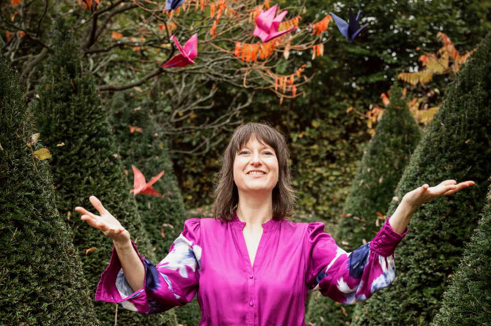 Tolk van Hartzaken Linda Blij staat tussen het groe in een paarse jurk. Zij lacht en gooit verschillende kleuren origami kraanvogels in de lucht.